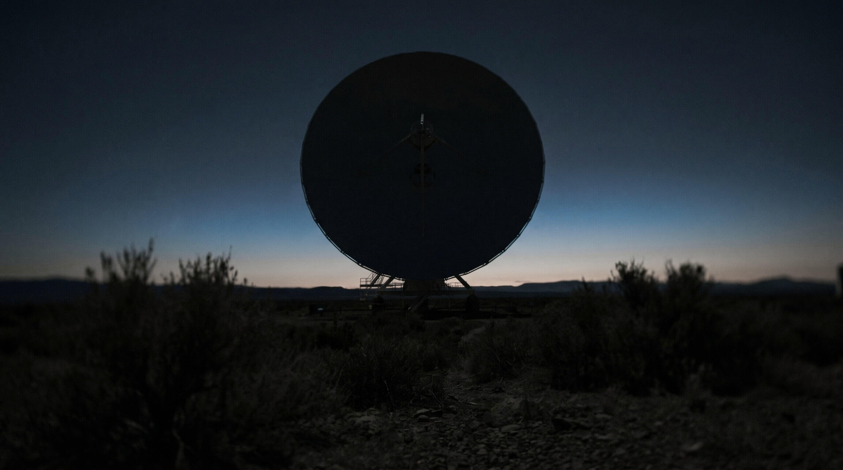 A large radio dish standing alone on a high desert plateau at dusk