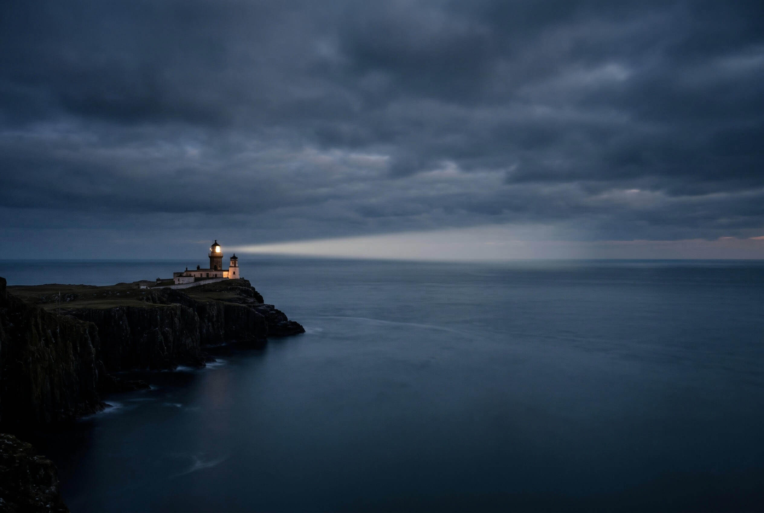 A lighthouse on a dark headland sweeping its beam across the sea at blue hour